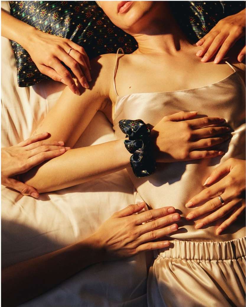 Overhead view of a person in satin pajamas lying in bed, surrounded by hands, creating a visually intimate and textured composition.