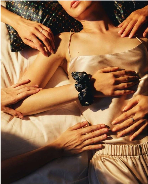 Overhead view of a person in satin pajamas lying in bed, surrounded by hands, creating a visually intimate and textured composition.