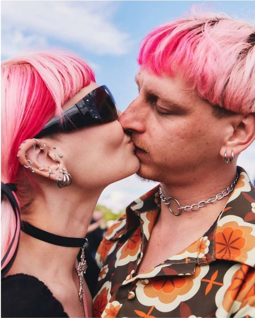 Close-up of a kissing couple with bright pink hair, silver chain jewelry, and vintage-style clothing outdoors on a bright, sunny day.
