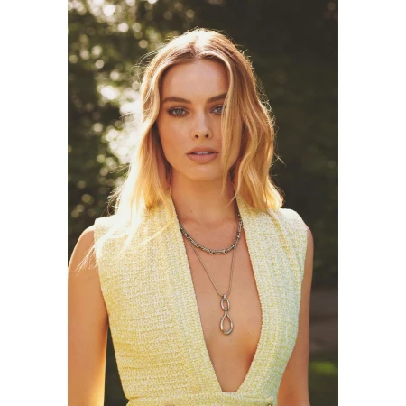 Close-up portrait of a blonde woman in a yellow tweed dress, gazing directly at the camera, with a blurred green leafy background.