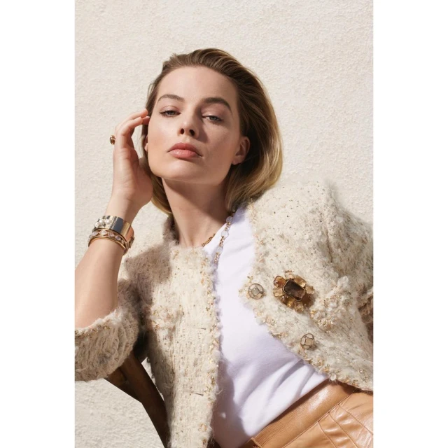 Blonde-haired woman, Margot Robbie, poses wearing a tweed jacket, gold bracelets, and tan leather skirt against a neutral backdrop.