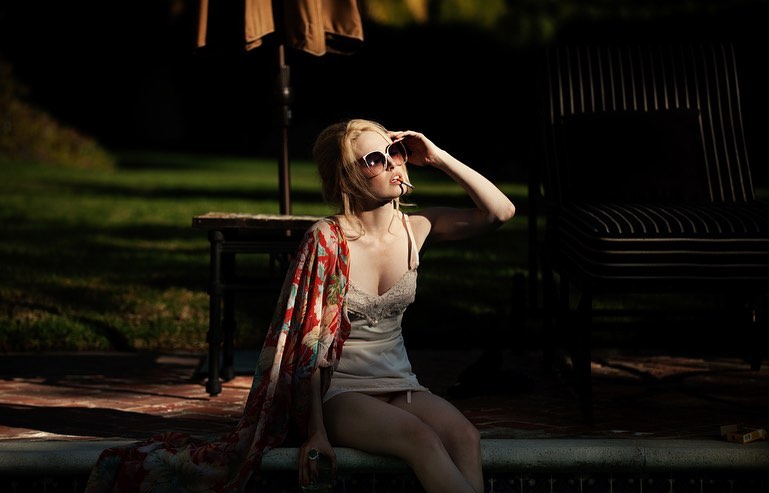 A blonde woman wearing sunglasses sits on the edge of a pool in dappled sunlight with a floral shawl draped over her shoulders.