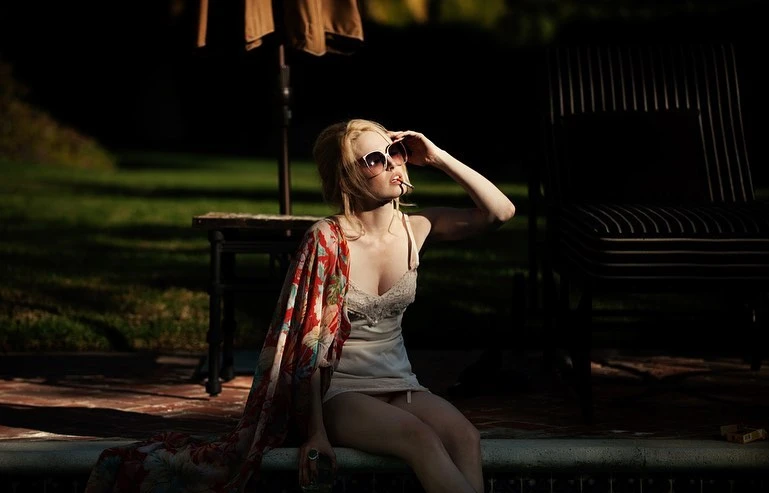A blonde woman wearing sunglasses sits on the edge of a pool in dappled sunlight with a floral shawl draped over her shoulders.