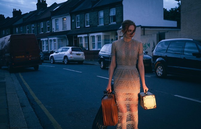 A woman in a gown stands on a city street holding luggage at dusk, flanked by parked cars and townhouses in the background.