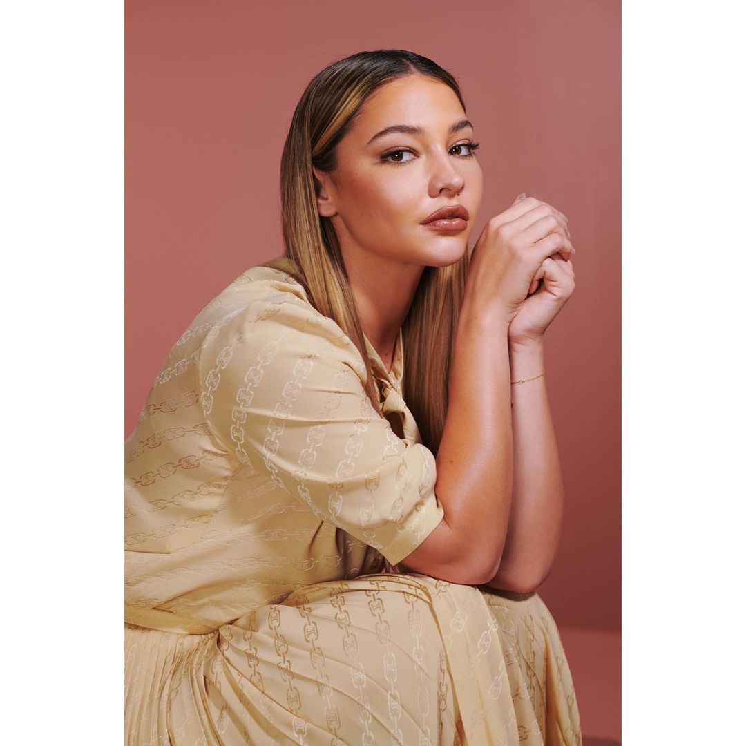 Portrait of Madelyn Cline in a beige dress with hands clasped, gazing at the camera against a rose-colored background in a photo studio.