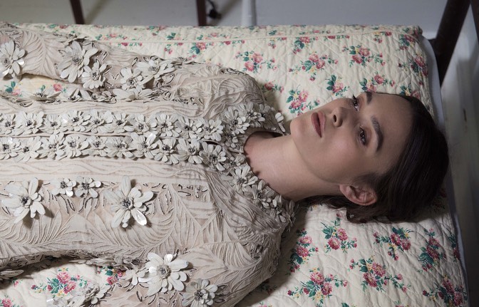 Woman in an embellished dress lying on a floral patterned bed with a dreamy, muted aesthetic and neutral color palette in a studio setting.