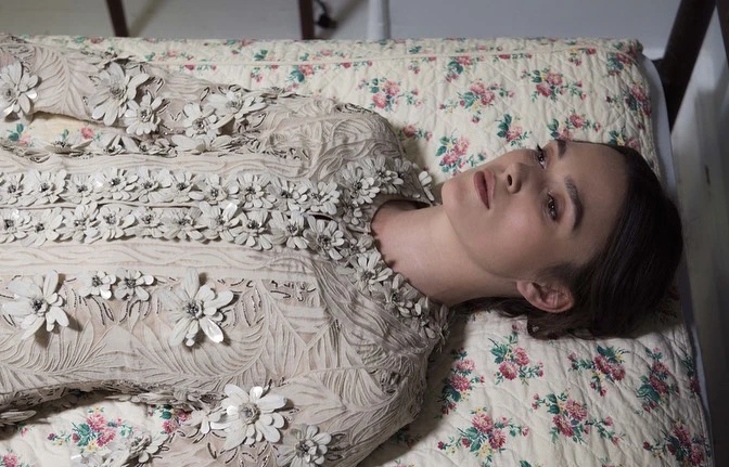 Woman in an embellished dress lying on a floral patterned bed with a dreamy, muted aesthetic and neutral color palette in a studio setting.