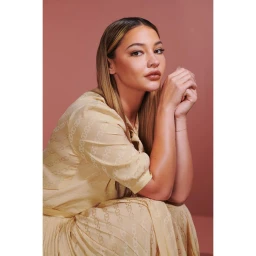 Portrait of a woman wearing a cream patterned dress with hands clasped together against a warm solid backdrop