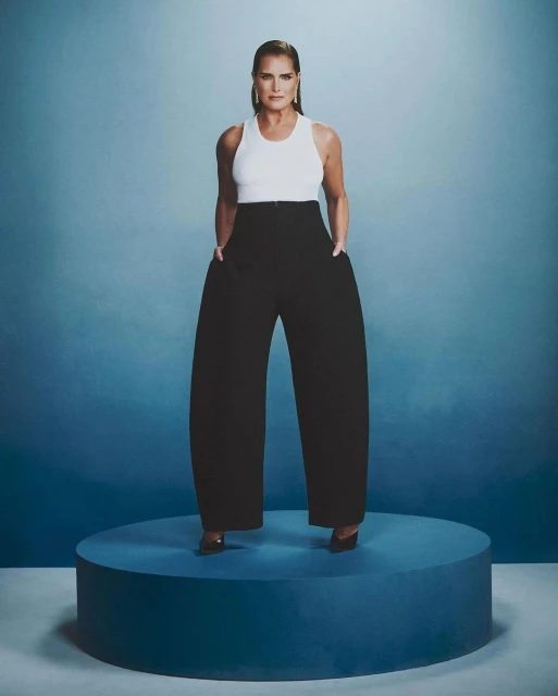 Studio shot of Brooke Shields in a white tank top and high-waisted black pants on a blue platform against a blue backdrop.