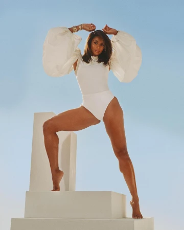 Woman in white bodysuit and blouse poses on a white block against a blue sky. She wears gold bracelets and has dark hair.