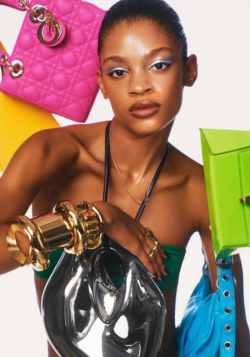 Portrait of a woman with colorful designer handbags, jewelry, and silver eyeshadow on a white backdrop in a fashion editorial studio shot.