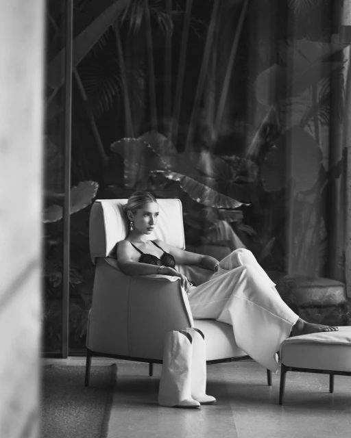 Black and white shot of a stylish adult female relaxing in a modern chair with cowboy boots at her feet inside an elegant home.