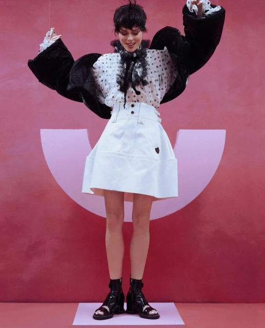 A woman in high-fashion, posing in a white skirt, polka dot blouse, and black jacket, against a pink backdrop with a logo.