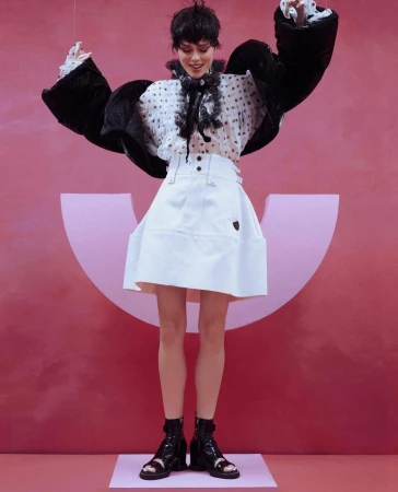 A woman in high-fashion, posing in a white skirt, polka dot blouse, and black jacket, against a pink backdrop with a logo.