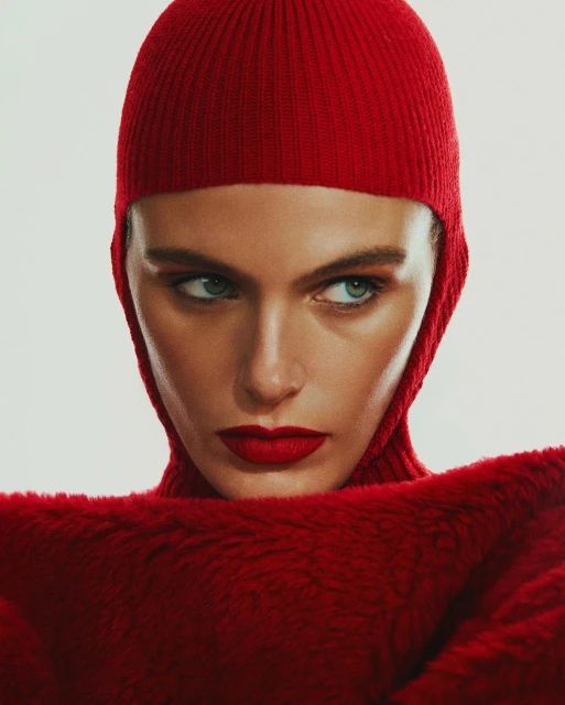 Close-up portrait of a woman with red lipstick wearing a matching red knit hat and faux fur sweater, against a white background.