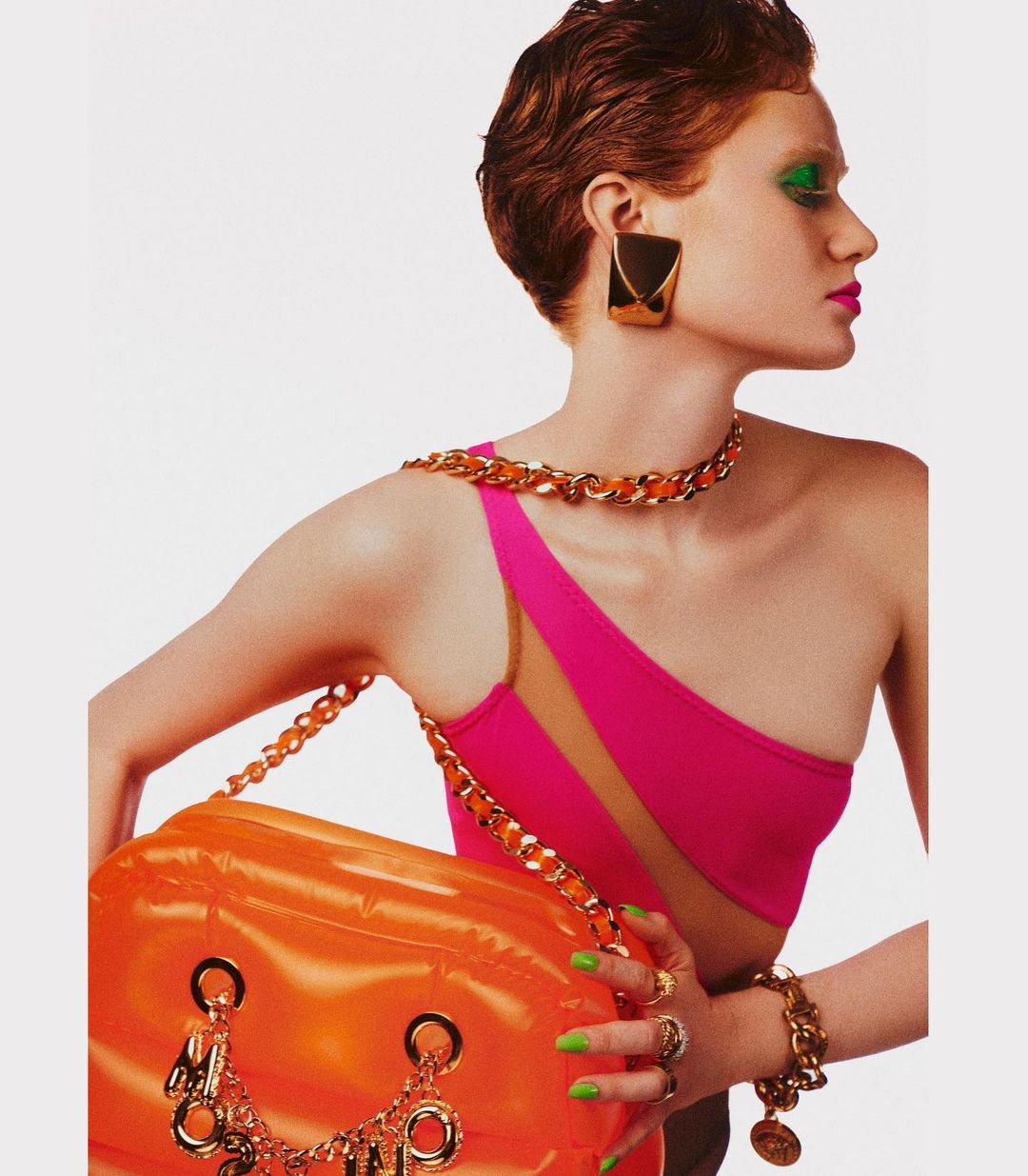 Fashion editorial shot of a red-haired woman with green eyeshadow, wearing a pink swimsuit, gold jewelry, and holding an orange bag.