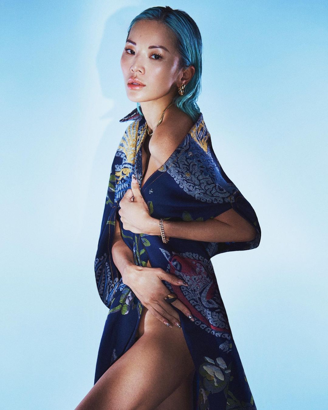 Attractive young adult woman with dyed blue hair poses in front of a light blue backdrop wearing a patterned blue dress.