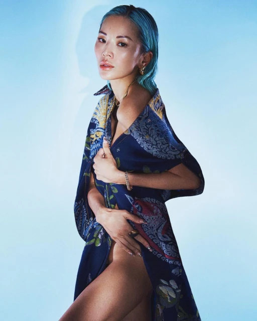 Attractive young adult woman with dyed blue hair poses in front of a light blue backdrop wearing a patterned blue dress.