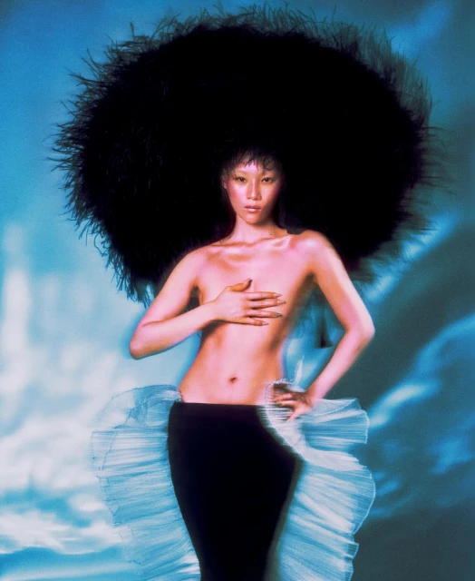 Striking studio shot of a woman with a large black feather headdress, standing in a black skirt and hand covering her chest.