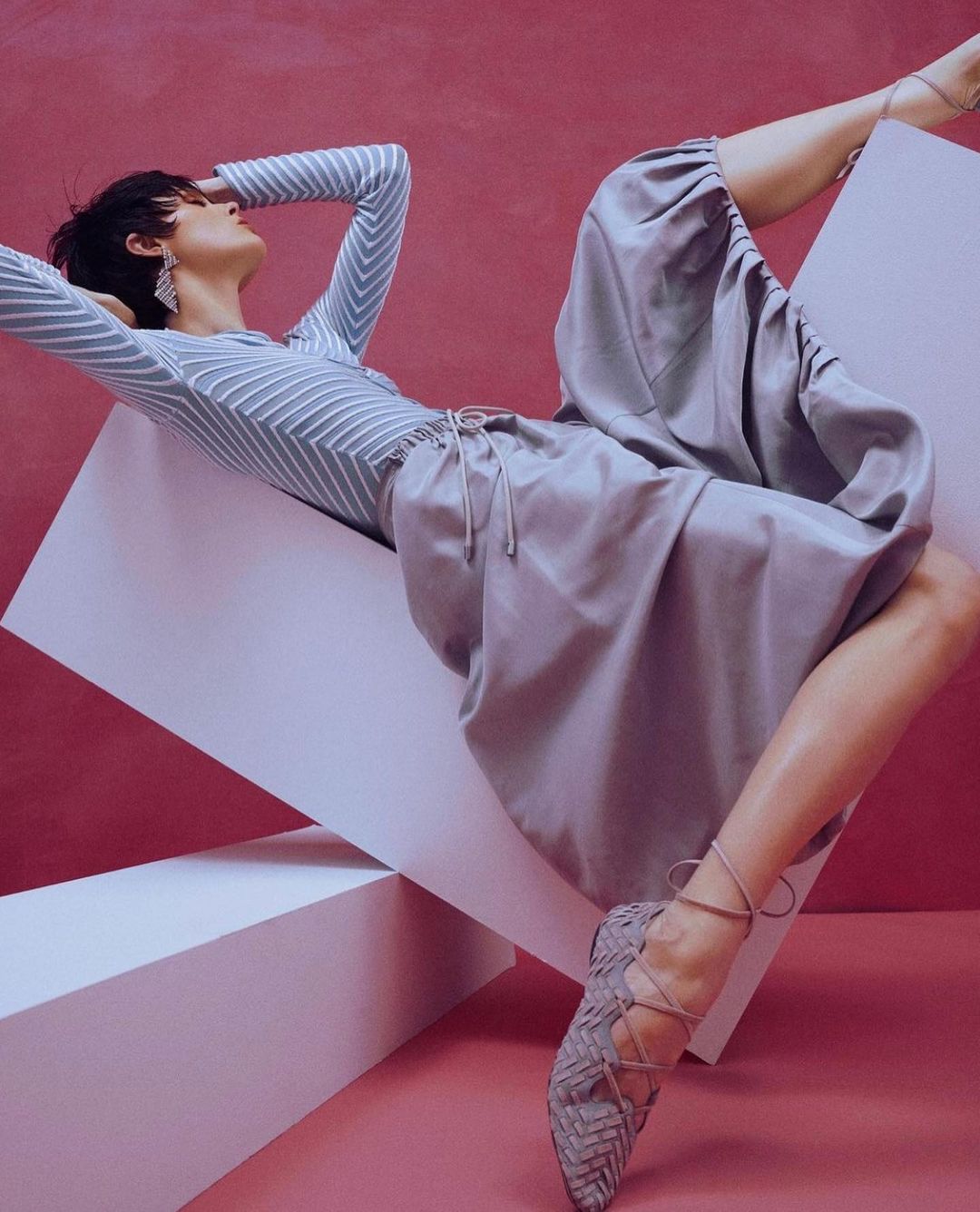 A woman lies among white geometric shapes in a fashion pose, wearing a striped top, a silk skirt, and woven ballet flats against a pink background.