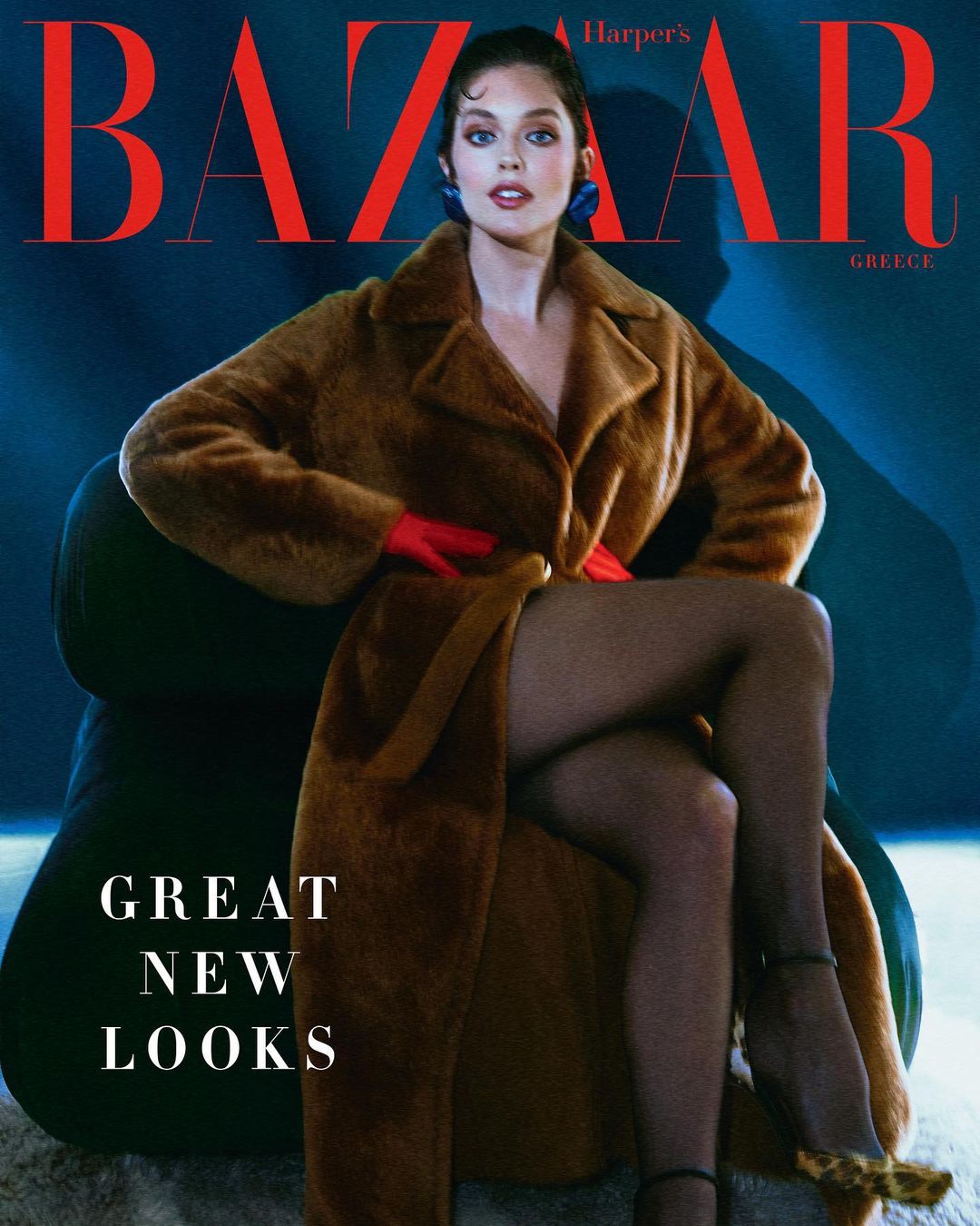 Magazine cover featuring a woman in a long brown coat sitting on an olive-green chair, set against a blue background with red text.
