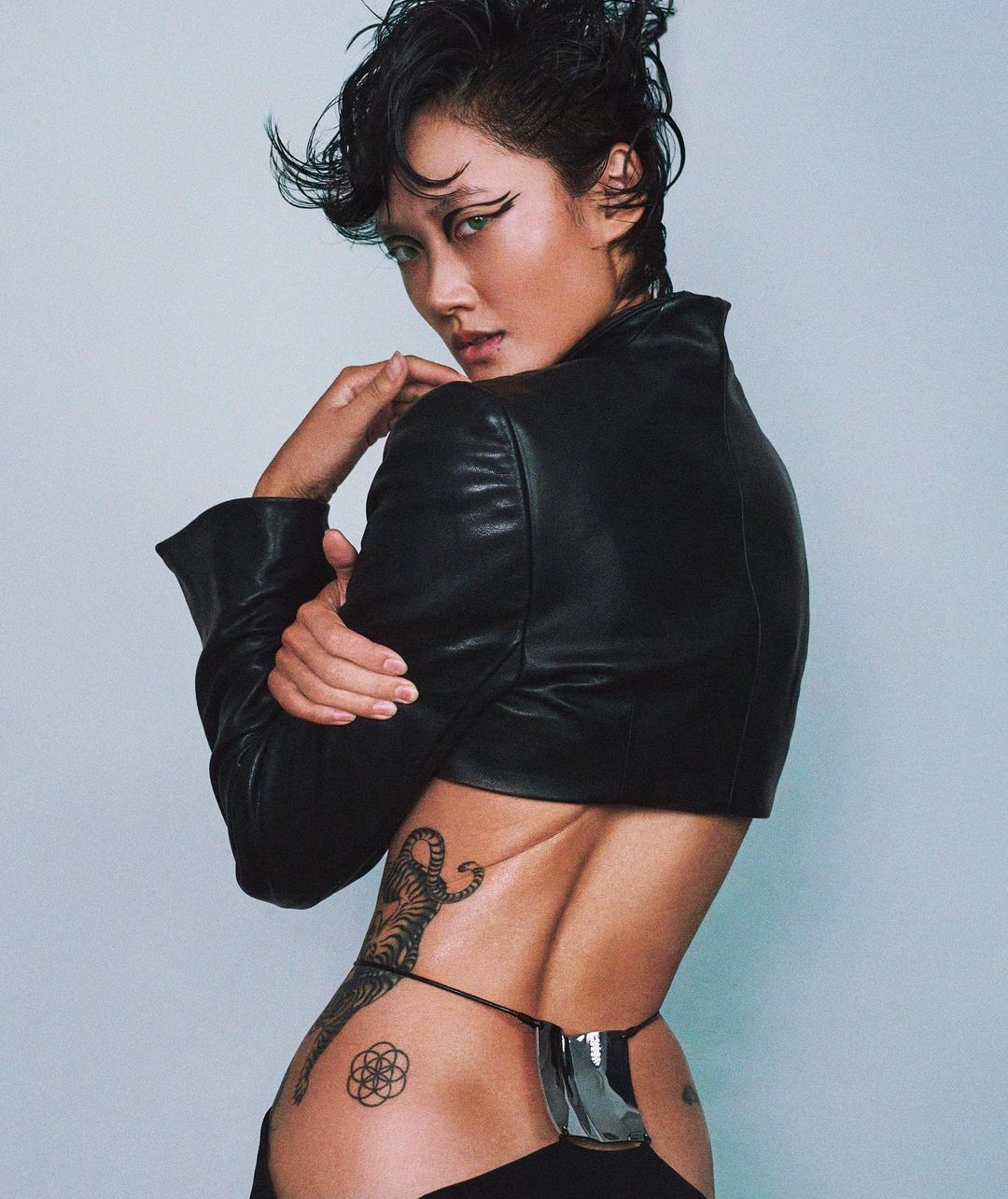 Striking portrait of an Asian woman with dark hair, sharp eyeliner, a black leather jacket, and distinctive tattoos on her lower back and hip.
