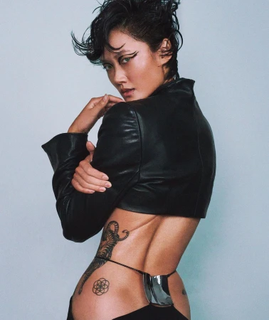 Woman in a black leather jacket with dark hair and eyeliner posing against a grey backdrop by Caleb & Gladys