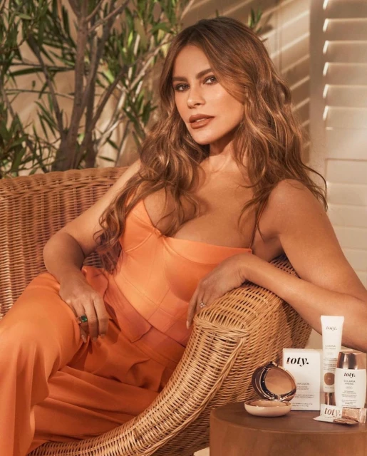 Sofia Vergara, smiling and seated in a wicker chair, models orange top and pants while showcasing beauty products in an indoor setting.