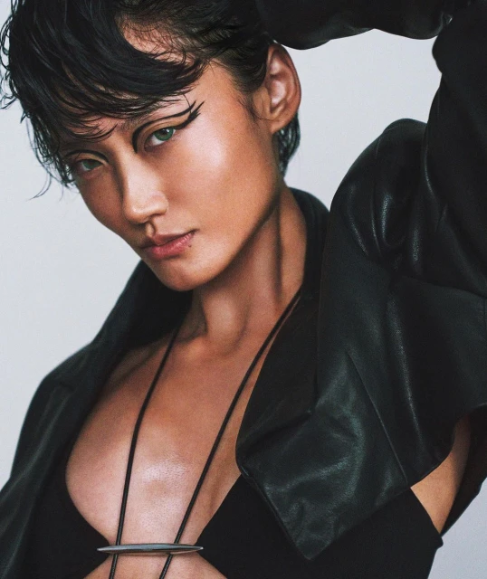 Close-up studio shot of a young adult woman in a black outfit and leather jacket, showcasing bold eye makeup and short, dark hair.
