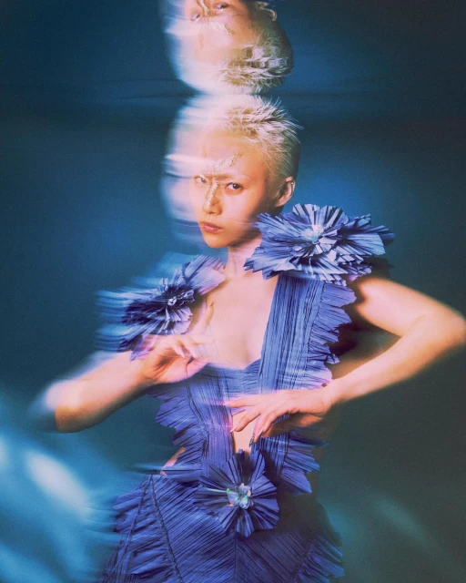 A woman with short blonde hair poses in a blue dress with floral accents in a blurred, dreamy composition with blue-toned lighting.