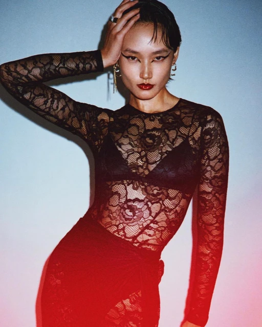 A woman with dark hair poses in black lace top and red lipstick, hand on her head, against a light blue backdrop, wearing statement earrings.