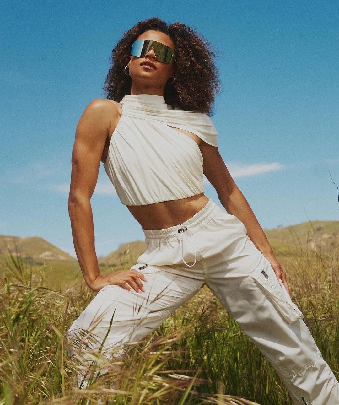 Woman in white crop top and cargo pants wearing reflective visor sunglasses outdoors in a grassy field under a clear blue sky