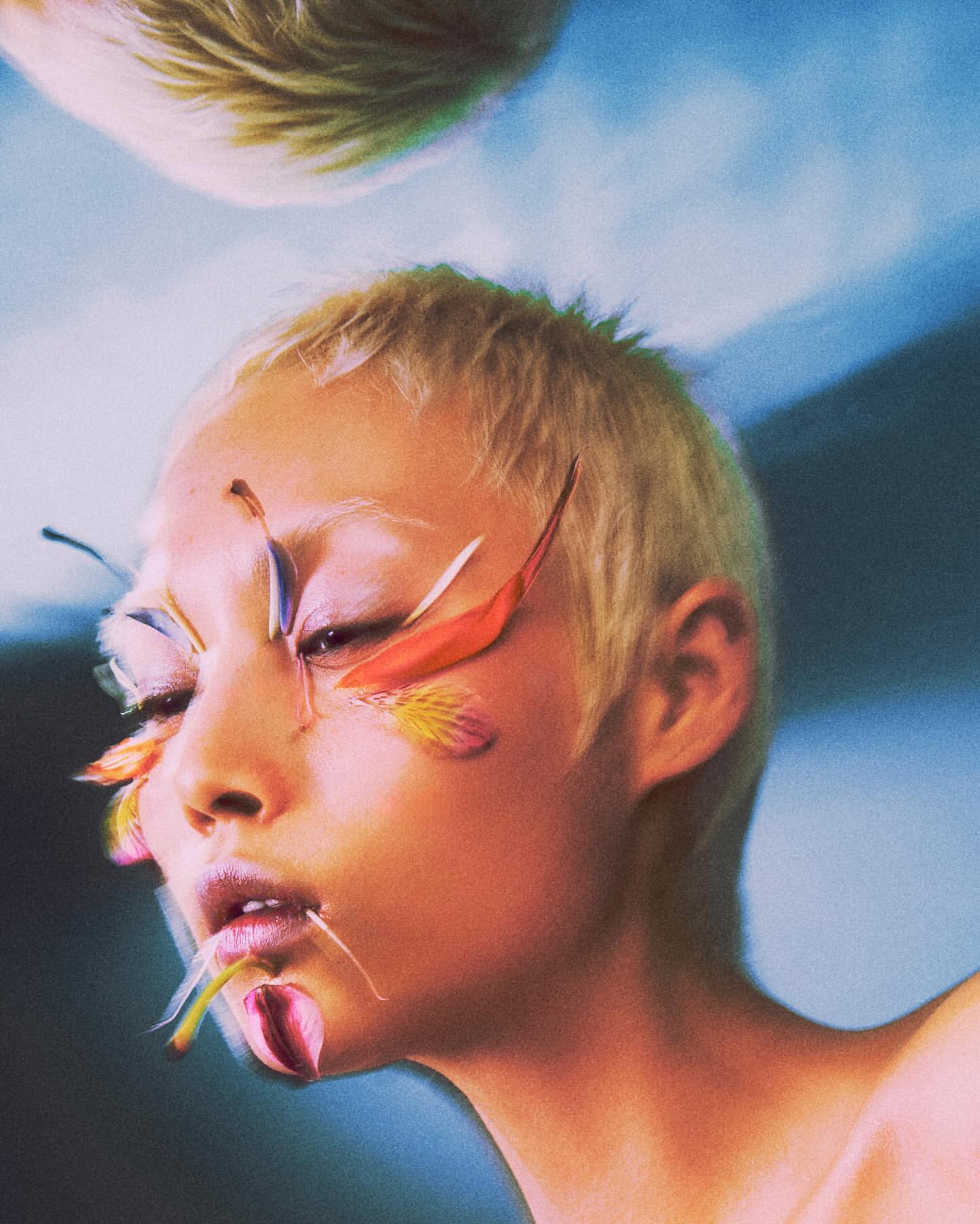 Artistic portrait of a woman with short hair, adorned with colorful flower petals on her face, against a blurred blue backdrop.