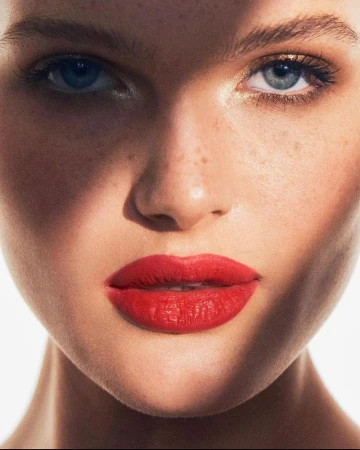 Close-up beauty portrait of a woman with vibrant red lipstick, light freckles, and striking blue eyes against a white backdrop.