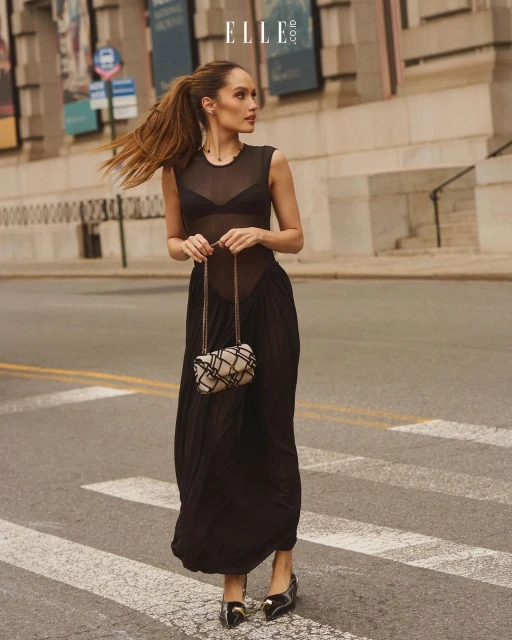 A woman in a black dress and carrying a bag walks across a crosswalk in an urban environment, with a large building in the background.