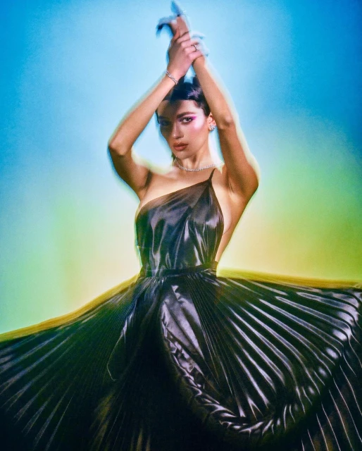 Elegant woman in a dark, flowing one-shoulder dress with a pleated skirt, hands raised, against a vibrant blue and yellow background.