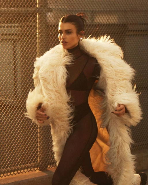 Woman in sheer black bodysuit and fuzzy white coat stands in front of chain link fence at sunset. Fashion editorial photography.
