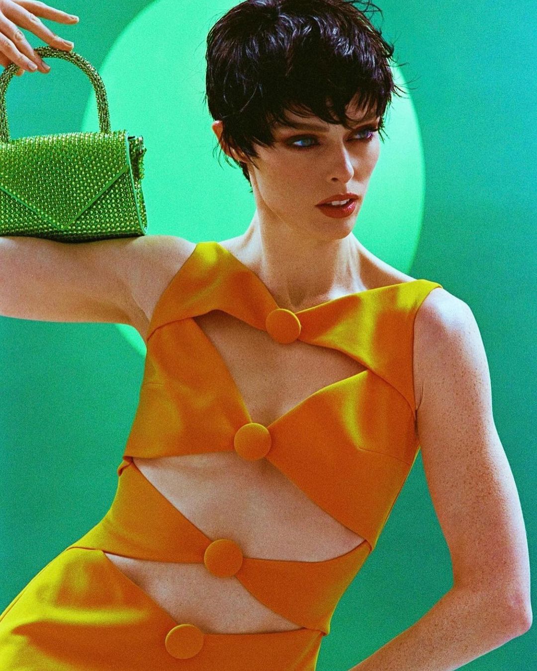 Stylish woman with short dark hair poses in an orange dress with cutouts, holding a green crystal handbag against a green backdrop.