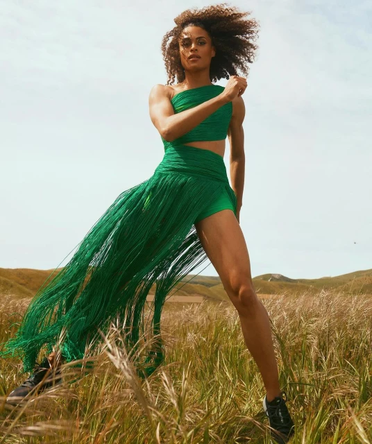 A curly-haired woman in a green dress strides through a golden field beneath a bright sky, captured in a fashion or athletic pose.