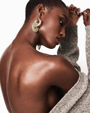 Close-up portrait of a Black model with short hair wearing large hoop earrings and a textured grey cardigan against a white background.