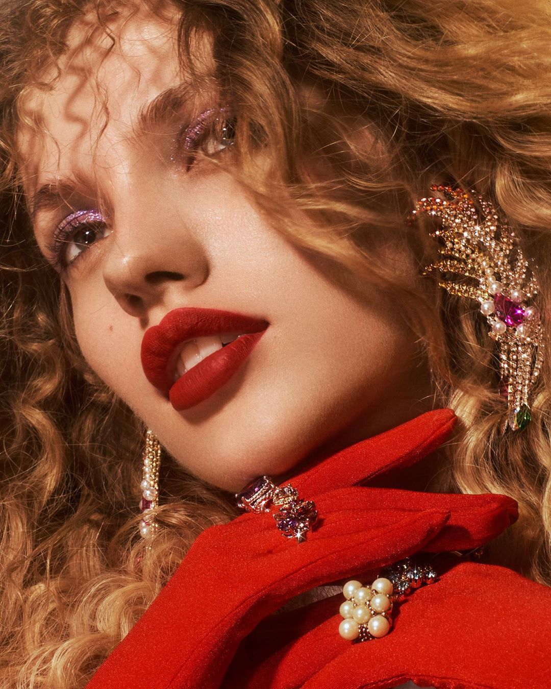 Portrait of a woman with red lipstick, wearing sparkling eye shadow and dangling jeweled earrings and rings and red gloves. Studio shot.