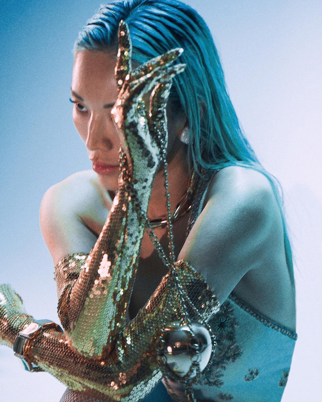 Elegant woman with blue hair and sequined gloves poses with a jeweled orb pendant against a dreamy light blue background.