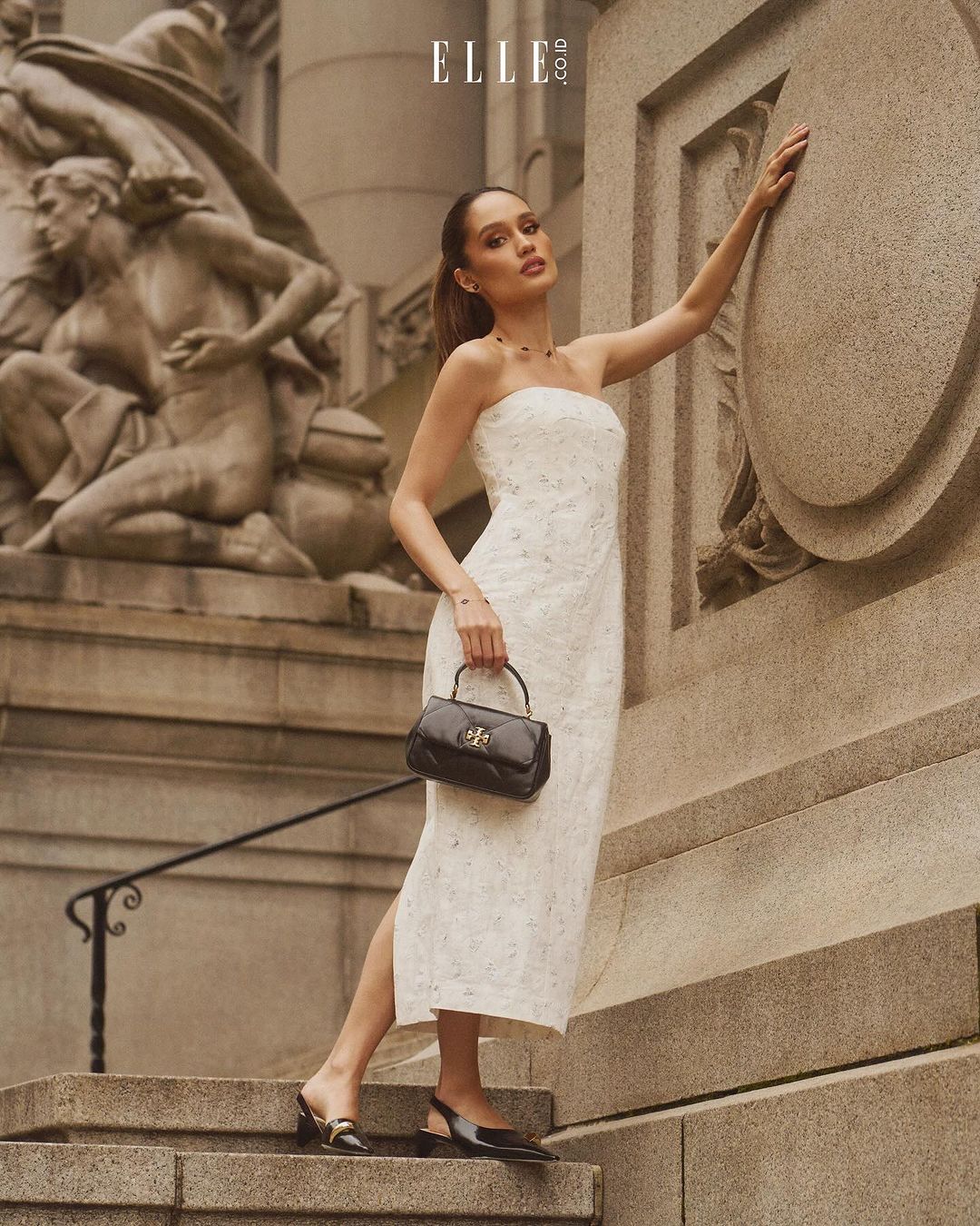 Elegant woman in a white dress poses near a marble statue and building, holding a black handbag, creating a classic outdoor fashion moment.
