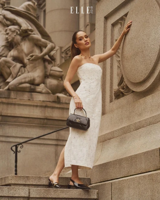 Elegant woman in a white dress poses near a marble statue and building, holding a black handbag, creating a classic outdoor fashion moment.
