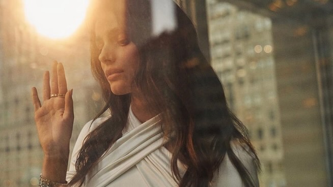 Woman in white top with raised hand showing ring, shot through glass with warm lens flare and city bokeh