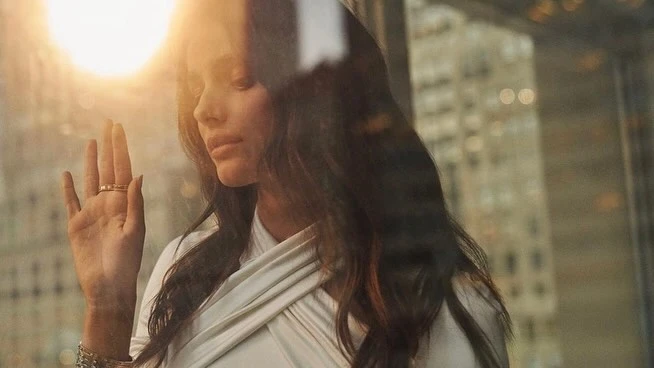 Woman in white top with raised hand showing ring, shot through glass with warm lens flare and city bokeh