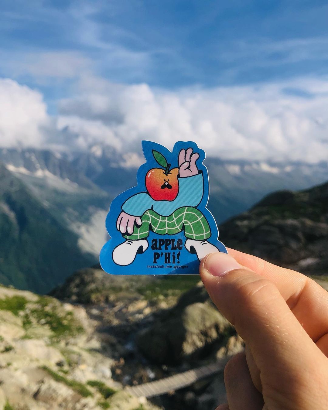 An apple cartoon sticker, 'Apple P'hi!' held by a person in front of a scenic mountain background on a clear day.