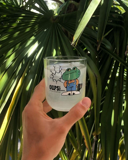 Hand holds a glass of cold beverage with a cartoon frog design and ice cubes; palm tree fronds visible in the background.