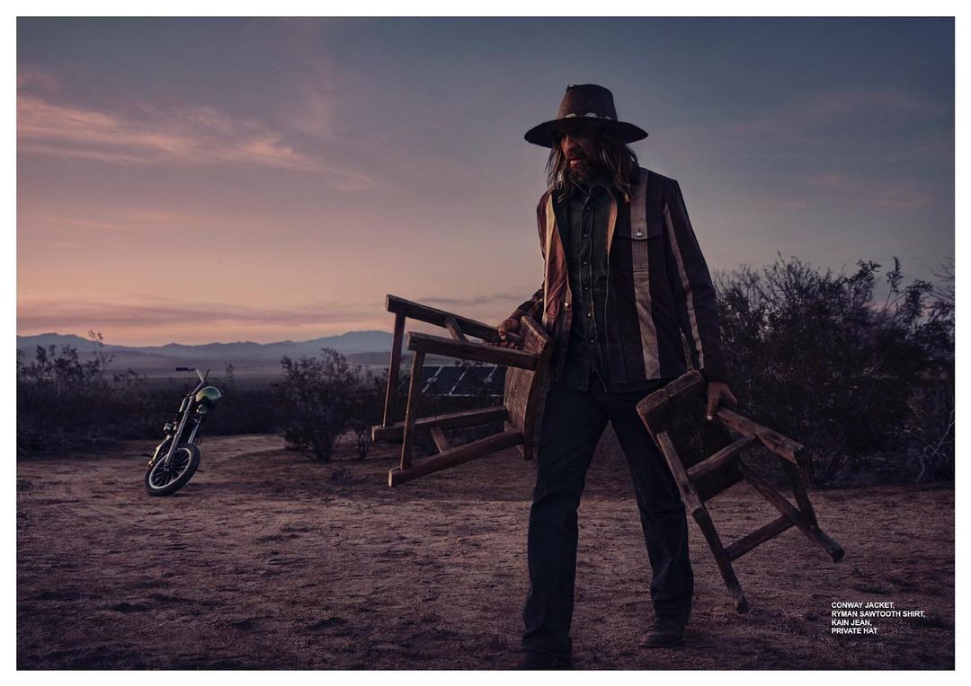 Adult male outdoors at dusk in cowboy hat and striped jacket, holding wooden chair parts, with a motorcycle on desert landscape.
