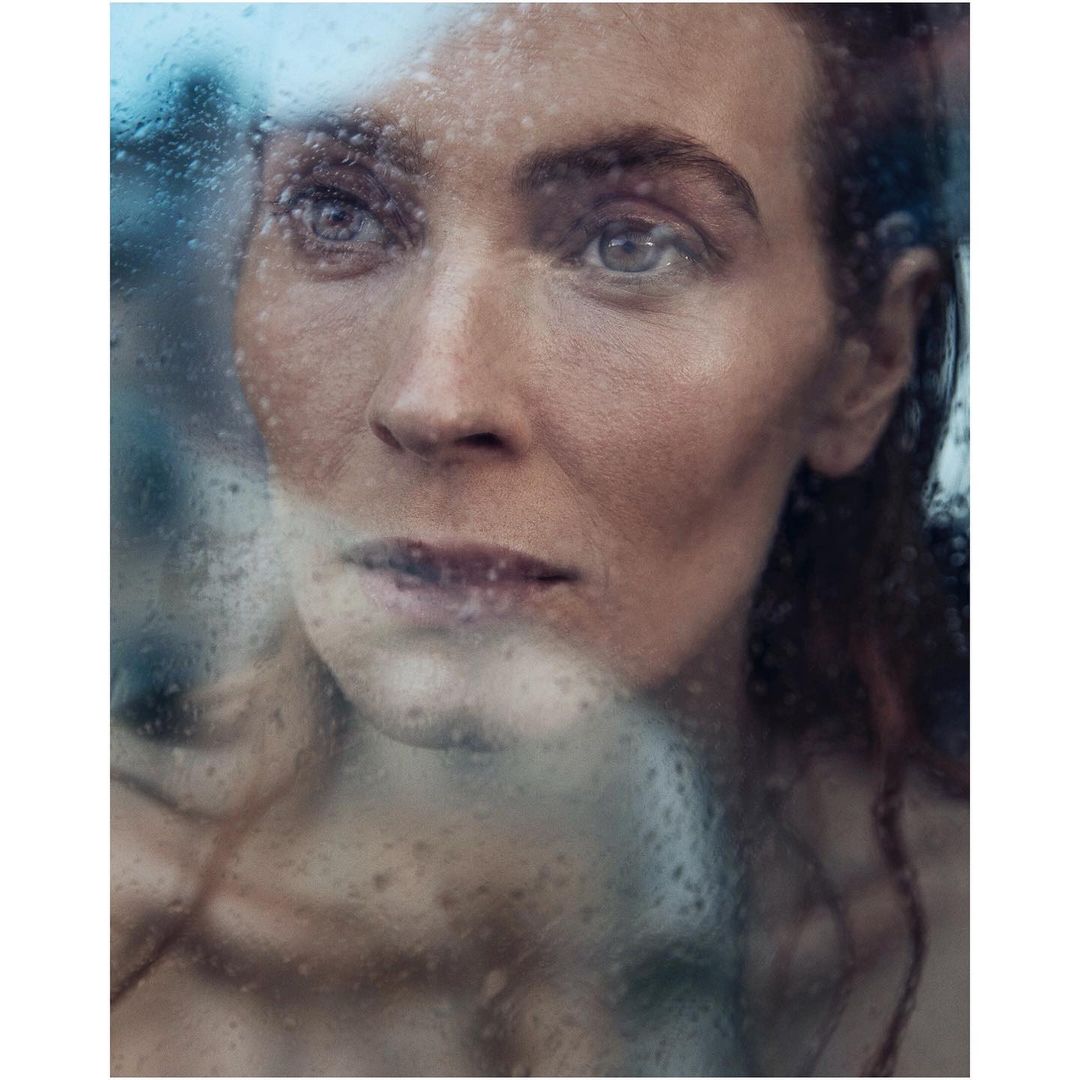 Close-up portrait of an adult woman with wet hair, looking pensively through a window covered in raindrops. Focus on her facial features.